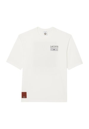 Camiseta blanca de manga corta con el texto "LACOSTE Roland Garros Club" y un gráfico de rejilla en el lado izquierdo del pecho, y un pequeño parche marrón con el logo del cocodrilo verde en el dobladillo.