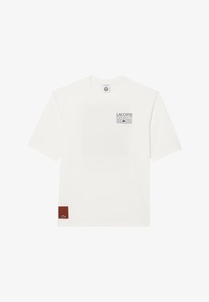 Camiseta blanca de manga corta con el texto "LACOSTE Roland Garros Club" y un gráfico de rejilla en el lado izquierdo del pecho, y un pequeño parche marrón con el logo del cocodrilo verde en el dobladillo.