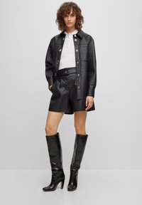 Czarna skórzana kurtka z teksturowanym wzorem, zestawiona z dopasowanymi szortami i czarnymi botkami do kolan. Biała koszula z kołnierzykiem pod spodem.