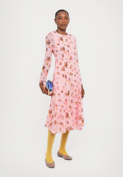 Roze bloemenjurk met lange mouwen, uitlopende zoom en een lichte stof. Gecombineerd met gele maillots en gestreepte schoenen, met een blauwe clutch in de hand.
