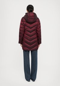 Vrouw in een lange bordeauxrode gewatteerde pufferjack met capuchon en wijde blauwe jeans, staand met de rug naar de camera op een witte achtergrond.