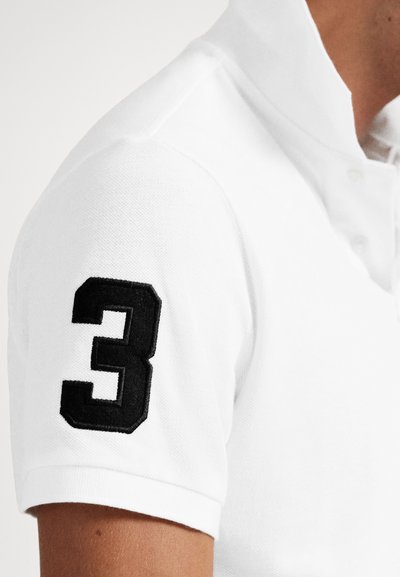 Polo branca com tecido texturizado e número "3" bordado a preto na manga esquerda. Detalhe da gola com botões visível.