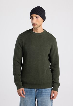 Man met een ribbelige donkergroene trui, lichtblauwe jeans en een zwarte gebreide muts, staande tegen een effen lichte achtergrond.
