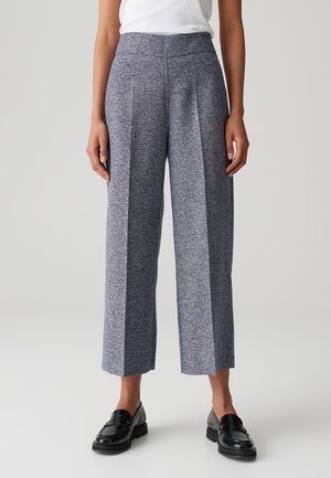 Mujer con pantalones grises de talle alto, pierna ancha y textura, y mocasines negros, de pie frente a un fondo claro y liso.