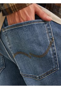 Jeans in denim con tasca posteriore caratterizzata da cuciture gialle visibili e un motivo a onde. Il materiale è morbido, con una tonalità blu scolorita e una texture sottile.