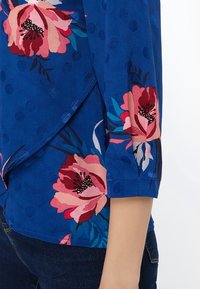 Blusa floreale blu con disegni di peonie rosa, maniche corte, tessuto testurizzato con pois e orlo inclinato.