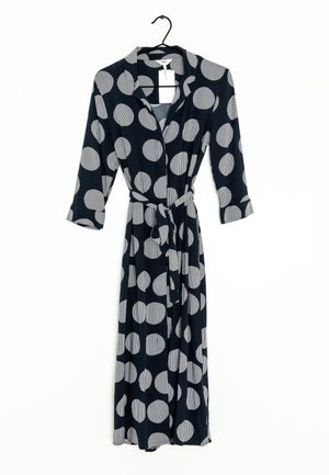 Langes schwarzes Kleid mit großen weißen Polka-Dots, langen Ärmeln, Kragen und Stoffgürtel, hängend an einem schwarzen Kleiderbügel vor weißem Hintergrund.