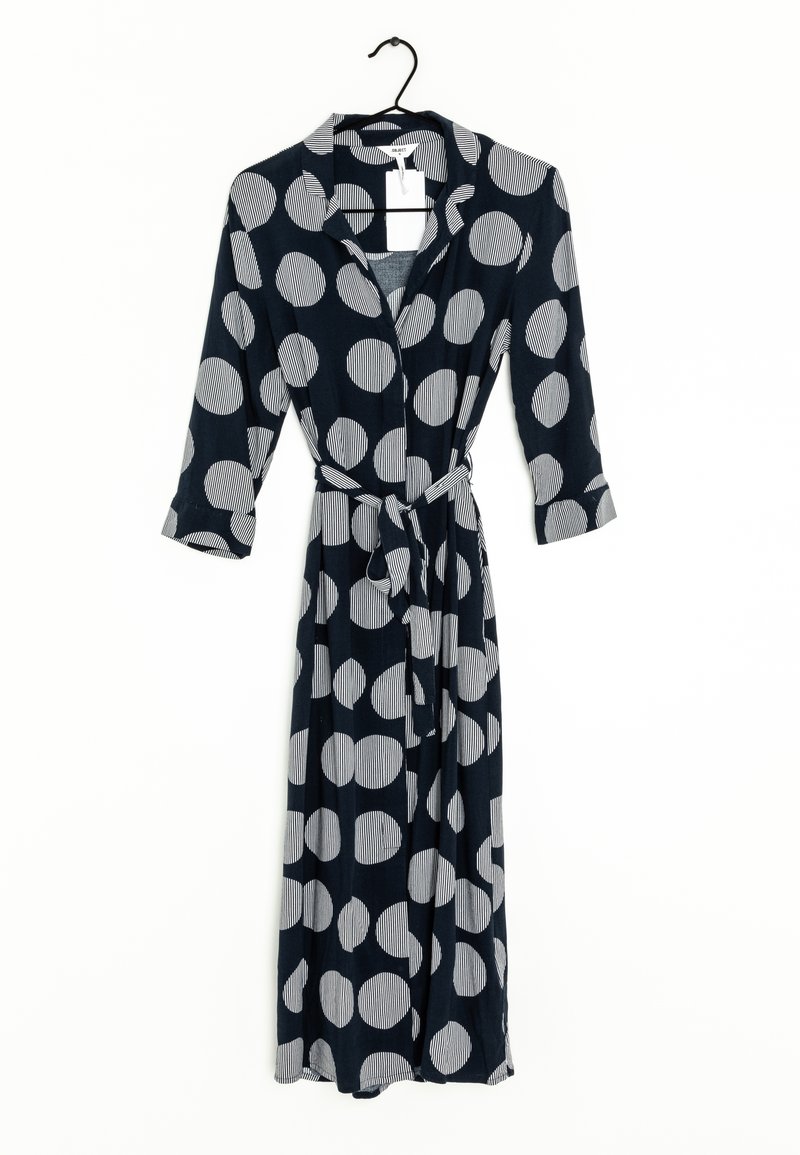 Longue robe noire à gros pois blancs, manches longues, col et ceinture en tissu, suspendue sur un cintre noir devant un fond blanc.