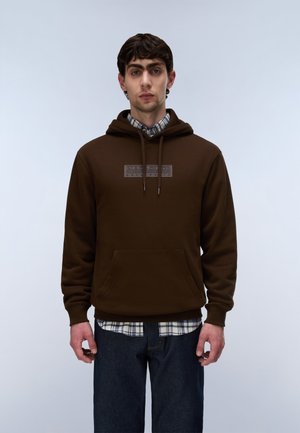Νέος άνδρας με σκούρα μαλλιά φοράει καφέ hoodie πάνω από ένα καρό πουκάμισο και μπλε τζιν, στέκεται μπροστά σε απλό φωτεινό φόντο.