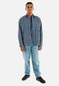 Camisa de botones en tela texturizada azul oscuro, llevada sobre una camiseta de color teal. Combinada con jeans azul claro y zapatillas negras con detalles marrones.