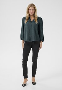 Blusa verde con texture, scollo a V e maniche a sbuffo, abbinata a pantaloni neri slim-fit e scarpe nere a punta.