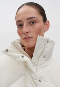 Weiße, gesteppten Puffermantel mit hohem Kragen, ausgestattet mit silbernen Druckknöpfen und einem weichen Innenfutter, das ein elegantes und minimalistisches Design zeigt.