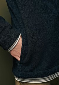 Schwarze Fleecejacke mit texturiertem Obermaterial, ausgestattet mit einer Seitentasche und gestreiften weißen und marineblauen Bündchen, die am Handgelenk sichtbar sind.