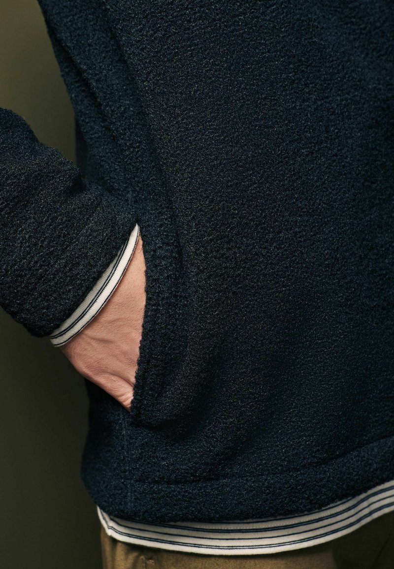 Schwarze Fleecejacke mit texturiertem Obermaterial, ausgestattet mit einer Seitentasche und gestreiften weißen und marineblauen Bündchen, die am Handgelenk sichtbar sind.
