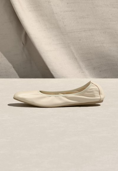 Μονό μπεζ δερμάτινο μπαλαρίνα παπούτσι με μαλακή στρογγυλεμένη μύτη, τοποθετημένο σε ανοιχτή επιφάνεια με υφασμάτινο μπεζ φόντο με υφή.