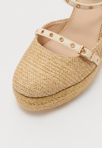 Espadrille beige con parte superior de paja trenzada, puntera redonda y correa para el tobillo con tachuelas en tono dorado y hebilla. Suela de plataforma de yute texturizada.