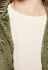 Olivově zelená bunda s lesklou, hladkou texturou, částečně rozepnutá přes krémové tričko s kulatým výstřihem z měkké látky.