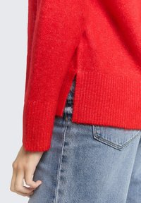 Rode gebreide trui met geribbelde cuffs en zijsplitten, gecombineerd met lichtblauwe denim jeans. De textuur lijkt zacht en warm.