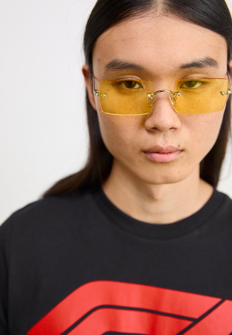 Ochelari de soare galbeni dreptunghiulari cu ramă metalică, având accente laterale unice, purtați peste un tricou negru cu un design grafic îndrăzneț roșu.