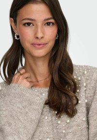 Beiger Strickpullover mit einer Auswahl an dekorativen Perlen, ausgestattet mit einem Rundhalsausschnitt und gerippten Bündchen. Das Haar ist lang und wellig.