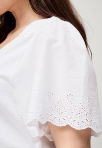 Blusa bianca con maniche a palloncino e delicato pizzo ad occhiello sull'orlo. Il materiale sembra leggero e strutturato.