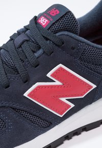 Gros plan sur une basket New Balance 565 noire avec un logo rouge et des lacets noirs sur fond blanc.
