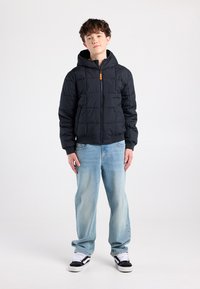 Zwarte donzen jas met een capuchon, ritssluiting, gewatteerd ontwerp en een oranje accent. Draagt het met lichtblauwe spijkerbroeken en zwarte sneakers.