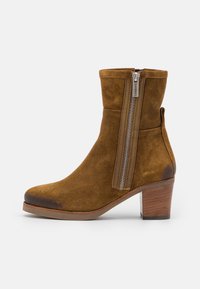 Bota de tobillo de ante marrón con punta redonda y cremallera lateral; presenta un tacón de madera apilada y una superficie lisa, ligeramente texturada.