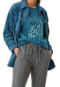 Camicia in velluto a coste blu sopra una felpa teal con testo grafico, abbinata a pantaloni grigi con cordino. Caratterizzata da trame morbide e vestibilità rilassata.