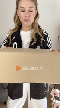 Caixa de cartão castanha com o logotipo laranja da Zalando visível, segurada à frente de uma pessoa que veste uma jaqueta preta com riscas brancas.