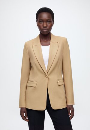 Femme portant un blazer beige ajusté sur un haut blanc et un pantalon noir, debout devant un fond blanc uni.