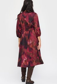 Lang burgunderfarget kjole med smocket midje, med en flytende silhuett og et flytende, abstrakt mønster i nyanser av burgunder og brunt.