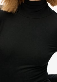 Blonde Person trägt ein enges schwarzes Rollkragenoberteil, von Hals bis Taille vor einem schlichten Hintergrund gezeigt.