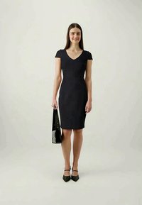 Mekko on musta, tiukka ja lyhythihainen, jossa on V-pään aukko. Malli pitää mustaa olkalaukkua ja on pukeutunut mustiin korkokenkiin.