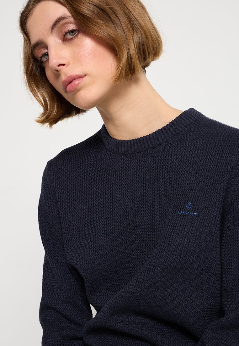 Granatowy sweter z dzianiny wykonany z teksturowanej tkaniny, z okrągłym dekoltem i małym akcentem logo w niebieskim kolorze na lewej klatce piersiowej.