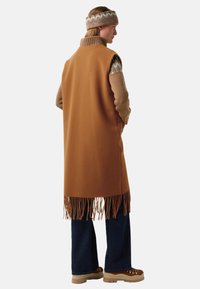 Braunwollweste mit Fransen, über einem beigefarbenen Strickpullover getragen. Dunkelblaue, ausgestellte Jeans und braune Schuhe runden den Look ab.