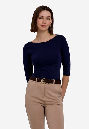 Mujer con pelo castaño hasta los hombros, con una camiseta azul marino de manga tres cuartos y pantalones beige con cinturón marrón, posando con las manos detrás de la espalda.