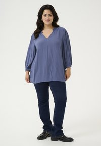 Blusa blu a maniche lunghe con scollatura a V e spalle arricciate, abbinata a jeans blu scuro e mocassini neri. Design semplice e privo di decorazioni.