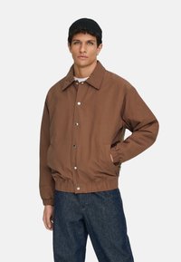 Braune Bomberjacke mit Knopfleiste, Hemdkragen, elastischen Bündchen und Bund. Kombiniert mit blauer Denim-Hose und einer schwarzen Mütze.