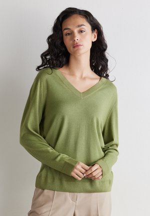 Frau mit langen, lockigen dunklen Haaren, die einen hellgrünen Pullover mit V-Ausschnitt und beige Hosen trägt, vor einem einfachen hellen Hintergrund stehend.