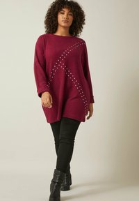 Maglione oversized bordeaux con scollo rotondo, decorato con borchie disposte in un motivo diagonale, abbinato a pantaloni neri aderenti e stivali neri.