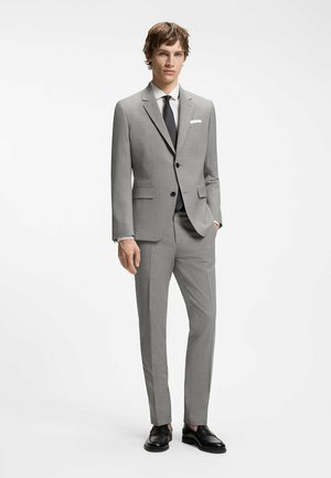 Joven de pie con un traje gris, corbata negra, camisa blanca, mocasines negros y una mano en el bolsillo sobre un fondo blanco.