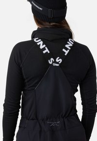 Sort ergonomisk overall med justerbare stropper, der har "SWEDEMOUNT" logo i hvid. Tekstureret sort langærmet trøje under.