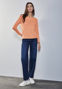 Pomarańczowa bluzka z długim rękawem i dekoltem boat, zestawiona z niebieskimi spodniami typu wide-leg i białymi trampkami, na neutralnym tle.