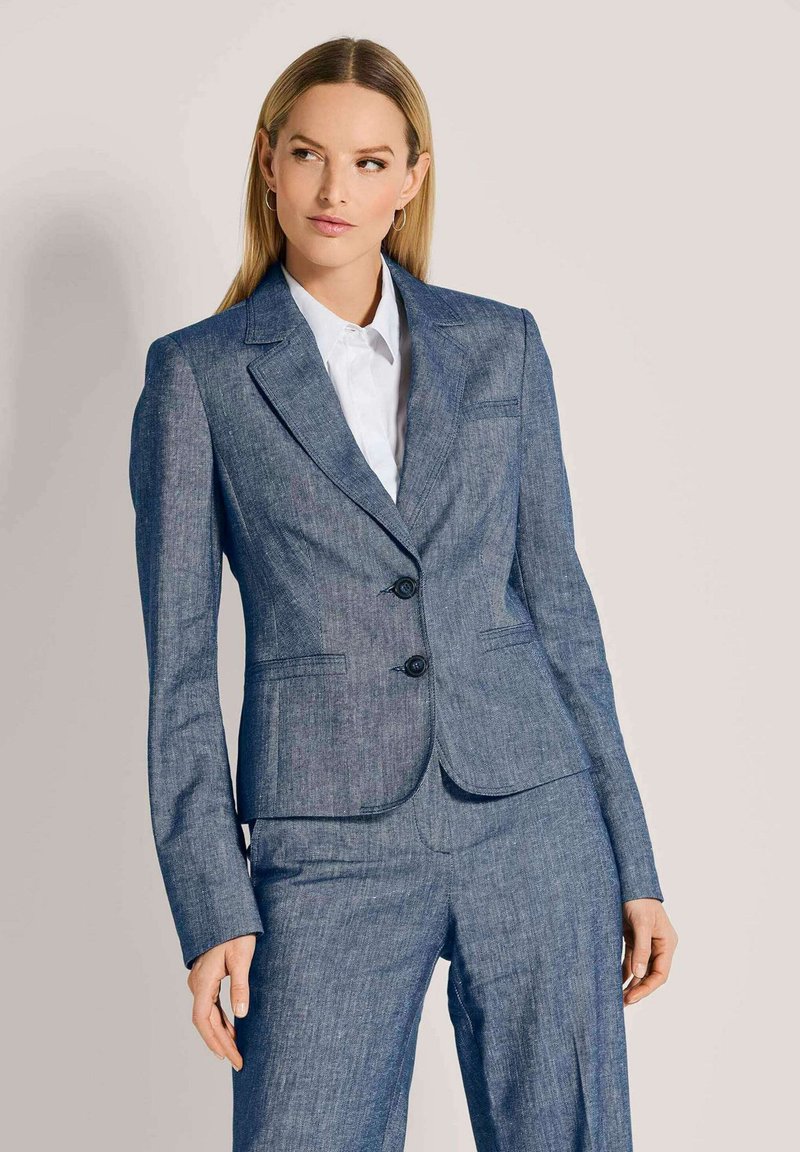 Vrouw met steil blond haar, gekleed in een strakblauw kostuum met een wit shirt, die iets naar rechts kijkt tegen een effen achtergrond.