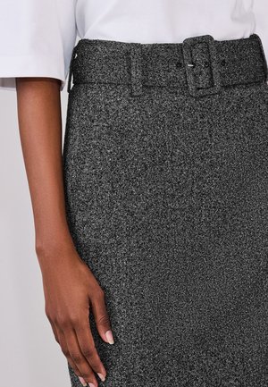 Hånd hviler ved siden af en grå fiskegratsmønstret nederdel med høj talje, bredt bælte og sølvspænde, kombineret med en hvid kortærmet top.