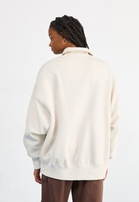 Oversized μπεζ φούτερ με ριμπ μανσέτες και στρίφωμα, που διαθέτει απλό καθαρό σχέδιο και απαλή υφή, παρουσιασμένο από την πίσω όψη.