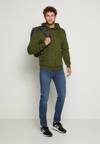 Grüne Zip-Hoodie mit Kordelzügen, zwei Vordertaschen, kombiniert mit blauen Jeans und schwarzen Sportschuhen. Ein grauer Rucksack hängt an einer Schulter.