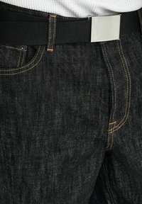 Tumši zili džinsu auduma bikses ar teksturētu pinumu, kontrastējošām dzeltens krāsas šuvēm un melnu austu jostu ar sudraba sprādzi.