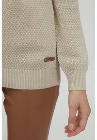 Beige strukturierter Pullover mit gerippten Bündchen, ausgestattet mit einem kleinen braunen Logoetikett. Kombiniert mit braunen Kunstlederhosen.